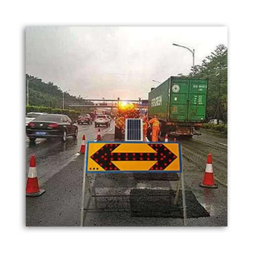 Traffic control sign with arrows indicating direction, solar panel on top, and construction workers nearby.