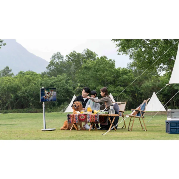 Family sitting outdoors with a dog, watching a television on a stand in a grassy area.